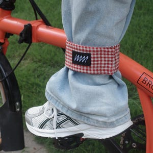 May include: A red and white gingham bicycle pant clip wrapped around the bottom of light blue denim jeans. The fabric band features a small black rectangular label with white zigzag line graphics. The clip is positioned on a leg near a bicycle pedal.