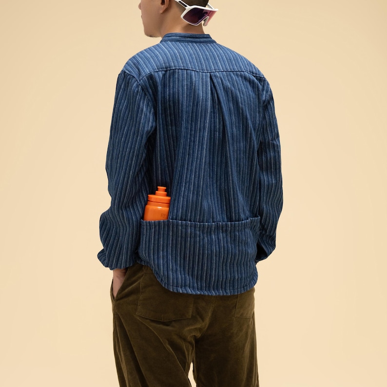 May include: A blue striped shirt with a large pocket, holding an orange water bottle. The shirt has long sleeves and a button-up design. The person is wearing brown pants and white sunglasses.