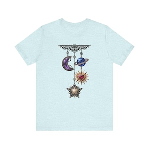 Puede incluir: Camiseta azul claro con un diseño celestial. El gráfico incluye una luna creciente morada, un planeta azul con anillos, un sol rojo en forma de corazón y un colgante de estrella. El diseño está suspendido de una filigrana negra decorativa.