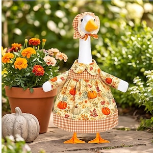 May include: A white goose figurine wearing a dress and bonnet featuring a pumpkin and autumn leaf pattern with orange gingham trim. The goose has a bright orange beak and feet, standing upright with arms extended outward in a static pose.