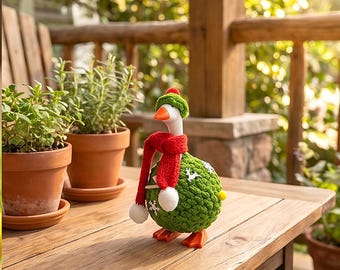 May include: A decorative goose figurine featuring a green crocheted body with white snowflake and reindeer patterns, wearing a red knit scarf with white pom-poms and a matching green hat. The goose has an orange beak and orange feet.