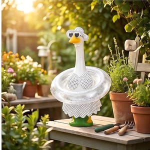 May include: A whimsical white goose figurine wearing a polka dot bikini, a matching sun hat with heart-shaped sunglasses, and a clear inflatable ring. The goose is on a green base, set in a garden scene.