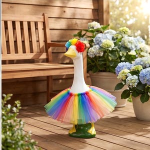 May include: A white goose statue wearing a vibrant rainbow-colored tulle tutu around its midsection and a matching multi-colored floral crown on its head. The goose has a yellow beak and yellow feet, standing upright as a decorative garden ornament.
