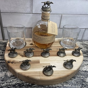 Puede incluir: Una botella de bourbon Blanton's con un tapón de caballo y jinete, dos vasos transparentes y siete figuras individuales de caballos y jinetes dispuestas sobre una tabla de madera semicircular con vetas de madera clara y oscura.