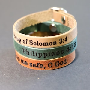 May include: Three stacked leather bracelets in tan, green, and brown, each engraved with a different Bible verse. The bracelets are fastened with a silver buckle. The text includes "Song of Solomon 3:4", "Philippians 4:13", and "Keep me safe, O God."