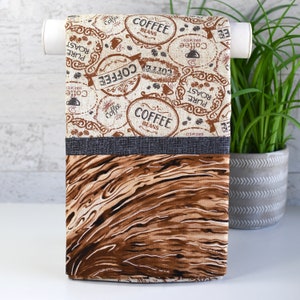 May include: A brown and beige kitchen towel with a woodgrain pattern on the bottom half. The top half features a repeating pattern of coffee bean illustrations and text that says "Coffee Beans", "Pure Roast", "Brewed", and "Coffee".