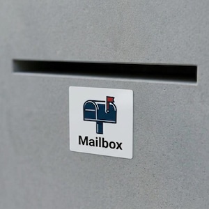 May include: A white square sticker with a blue mailbox illustration and the word "Mailbox" in black text. The mailbox has a red flag. The sticker is affixed to a gray concrete surface.