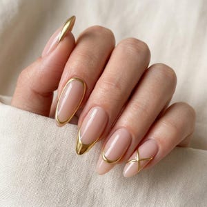 May include: Oval-shaped fingernails with a glossy nude base colour, featuring metallic gold accents. The designs include gold-rimmed edges, a solid gold tip, and a gold cross-shaped detail on the smallest nail, creating a polished manicure look.