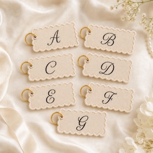 May include: Seven rectangular beige fabric tags with scalloped edges, each featuring a black embroidered letter from A to G in a cursive script. Each tag is attached to a small gold-toned metal ring for hanging or attachment purposes.
