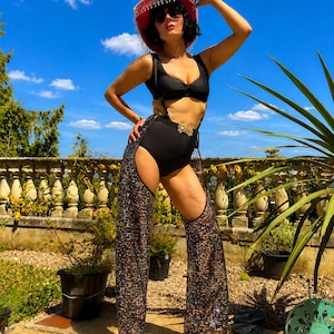 May include: A black bikini top and high-waisted bottoms paired with sparkling, multi-colored sequined chaps. The outfit includes a pink cowboy hat with dangling silver fringe, a gold butterfly belt buckle, and dark sunglasses under a blue sky.