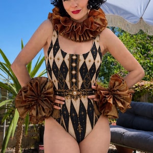 May include: A one-piece swimsuit featuring a black and cream diamond harlequin pattern with gold star accents, a central vertical panel with pearl-like details, and large, voluminous bronze-colored ruffled fabric accents at the neck and wrists.