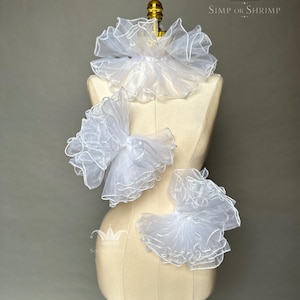 Puede incluir: Tres volantes de organza blanca transparente con bordes acabados expuestos en un maniquí. Los volantes presentan un diseño voluminoso y en capas con costuras delicadas. El texto SIMP OR SHRIMP con un icono de corona es visible arriba.