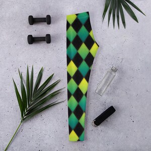 May include: A pair of leggings with a harlequin pattern in black, green, and yellow. The leggings are displayed on a gray surface with two black dumbbells, a clear water bottle, and a black wristband. Green palm leaves are also present.