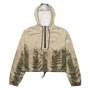 Puede incluir: Una chaqueta cortavientos corta de color beige con un estampado de hojas de helecho en verde oscuro. La prenda incluye una capucha con forro interior blanco, una cremallera frontal negra a media altura con cordones negros y un dobladillo ajustable.