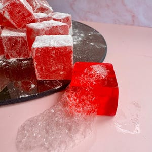 Puede incluir: Primer plano de dulces turcos rojos espolvoreados con azúcar glas. Una pieza está aislada, con una textura burbujeante y translúcida. Los dulces están sobre un plato negro sobre un fondo rosa.