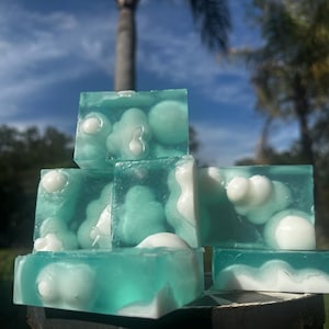 May include: Six turquoise soap bars with white, cloud-like shapes inside, stacked in a pyramid. The background shows a blue sky and palm trees, creating a natural, spa-like aesthetic.