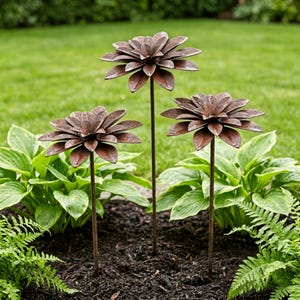 May include: Three metal flower garden stakes with a rustic brown finish. Each flower has multiple layered petals and is mounted on a slender stem. The stakes are placed in a garden bed with green foliage and grass in the background.