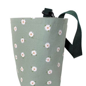 May include: A sage green fabric bag with a white daisy pattern. The bag has a dark green strap and a black clip. The bag is cylindrical and appears to be made of a sturdy fabric.
