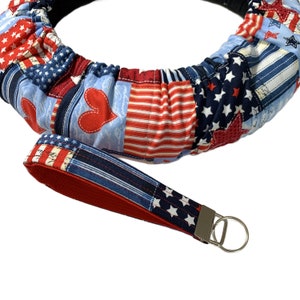 May include: Red, white, and blue patterned steering wheel cover with a matching key chain. The cover is made of fabric and has a ruffled design. The key chain has a silver ring.