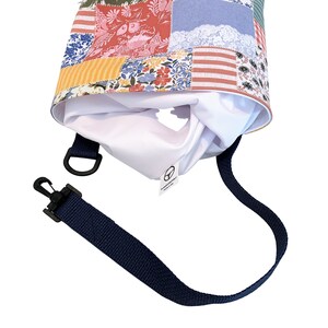 K&ouml;nnte beinhalten: Eine wei&szlig;e Tragetasche mit Patchwork-Design, das verschiedene florale Muster und Farben wie Rot, Blau, Gelb und Gr&uuml;n aufweist. Die Tasche hat einen verstellbaren dunkelblauen Trageriemen mit einem Metallclip.