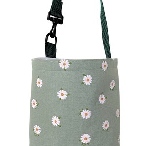 May include: A sage green tote bag with a daisy pattern. The bag has a dark green strap and a black clip. The daisies are white with yellow centers. The bag is designed for carrying items.