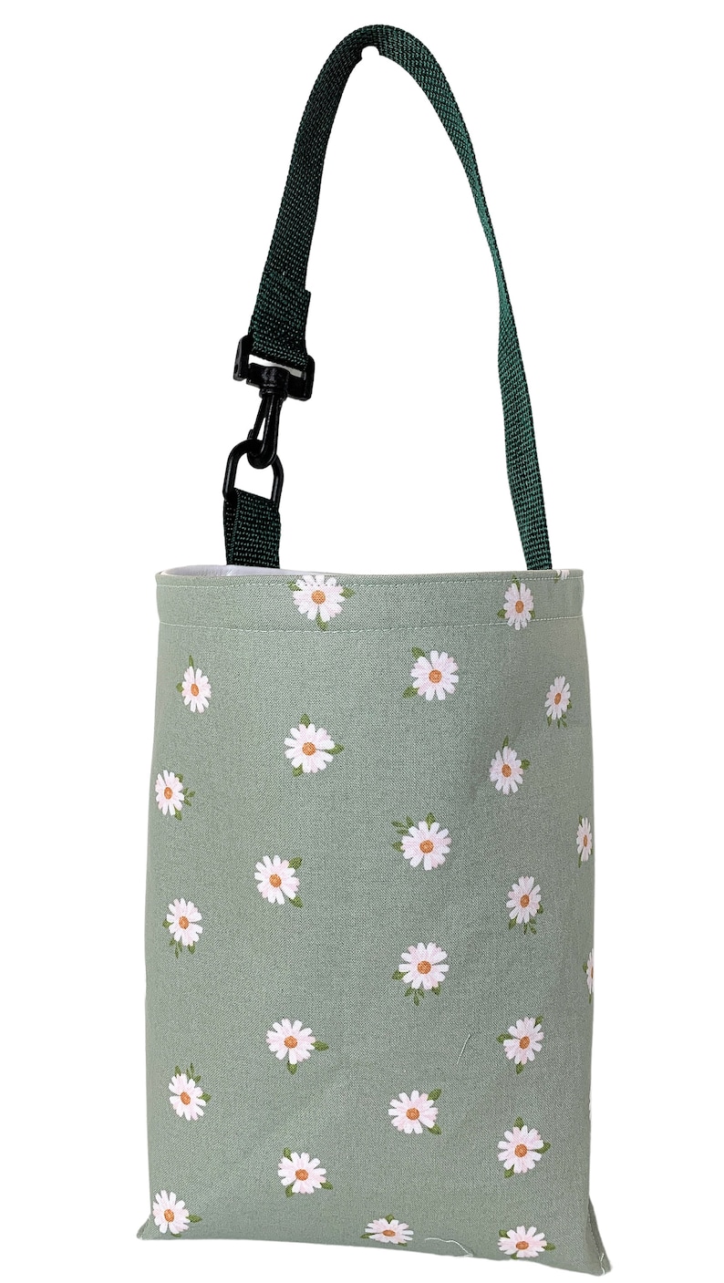 May include: A sage green tote bag with a daisy pattern. The bag has a dark green strap and a black clip. The daisies are white with yellow centers. The bag is designed for carrying items.