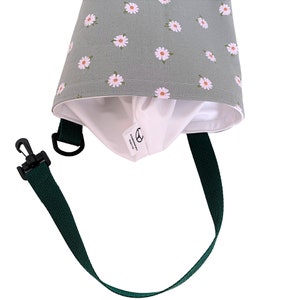 May include: A green fabric bag with a daisy pattern and a white interior. The bag has a dark green strap and a black clip. The bag's interior has a white tag with a logo and text.
