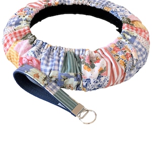 May include: A patchwork steering wheel cover with a matching wristlet keychain. The cover features a variety of fabric patterns, including floral, gingham, and striped designs in shades of red, pink, blue, and yellow. The wristlet has a blue fabric base with a floral pattern.