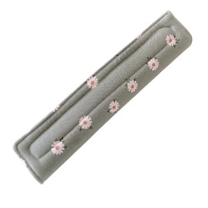 May include: A gray padded shoulder strap cover with a daisy floral pattern. The cover is rectangular with rounded ends and features a repeating pattern of white daisies with pink centers.