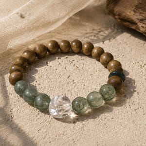 Puede incluir: Una pulsera de cuentas con una mezcla de cuentas de piedra natural. La pulsera incluye cuentas redondas de madera marrón claro, cuentas de piedra verde-gris y una cuenta de piedra clara tallada. La pulsera se exhibe sobre una superficie texturizada de color claro.