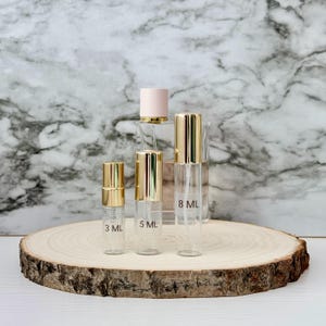 Pode incluir: Frascos de perfume de vidro transparente com tampas douradas, rotulados 3 ml, 5 ml e 8 ml, são exibidos em uma fatia de madeira. Um frasco de perfume maior com tampa rosa está no fundo. O fundo é um padrão de mármore.