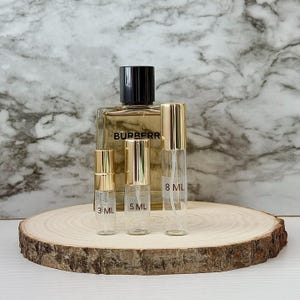 Puede incluir: Un frasco de perfume y tres frascos de spray de vidrio con tapas doradas están colocados sobre una rodaja de madera. El frasco de perfume tiene una tapa negra y la palabra "BURBERR" está impresa en la parte delantera. Los frascos de spray están etiquetados 3 ML, 5 ML y 8 ML.