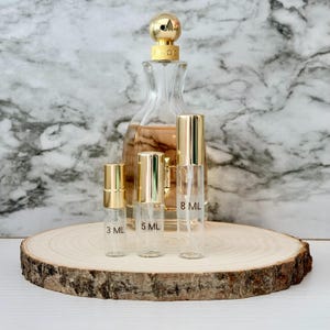 Puede incluir: Colección de frascos de perfume y atomizadores sobre una rodaja de madera. El conjunto incluye un frasco de perfume grande con tapa dorada y varios atomizadores de vidrio más pequeños etiquetados 3 ml, 5 ml y 8 ml, todos con tapas doradas.