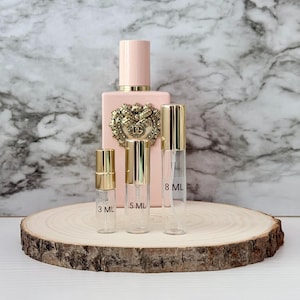 Puede incluir: Un frasco de perfume rosa con un emblema dorado y una tapa dorada, acompañado de tres frascos de muestra de perfume de vidrio transparente con tapas doradas. Los frascos de muestra están etiquetados con 3 ml, 5 ml y 8 ml. Los artículos se exhiben sobre una rodaja de madera.