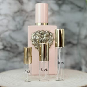 May include: A light pink perfume bottle with a gold emblem and a matching pink cap. Three clear glass spray bottles with gold caps are in front, labeled 3 ML, 5 ML, and 10 ML. The bottles sit on a wooden slice.