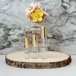 Pode incluir: Um frasco de perfume transparente com uma tampa em forma de margarida, juntamente com três frascos de spray recarregáveis. A tampa de margarida apresenta pétalas rosa, brancas e amarelas com detalhes dourados. Os frascos recarregáveis são rotulados com 3 ml, 5 ml e 8 ml.