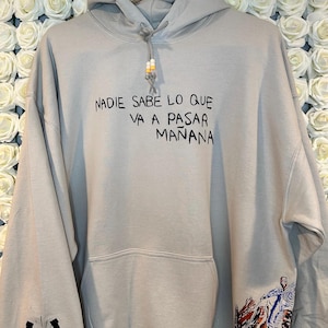 May include: A light beige hoodie with a drawstring hood and a front pocket. The text "NADIE SABE LO QUE VA A PASAR MANANA" is printed in black on the front. The left sleeve has a horseshoe and eye graphic, and the right sleeve has a colorful illustration.