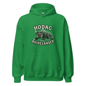 May include: A bright green hooded sweatshirt featuring a graphic of a green, horned, sharp-toothed creature. The text HODAG is printed in an arched cream font above the creature, and the word RHINELANDER is printed in a matching font below it.
