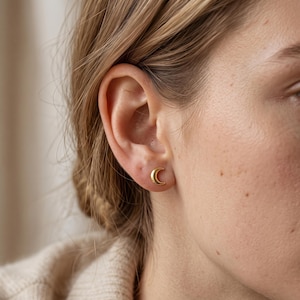 May include: A small, polished gold-tone stud earring shaped like a crescent moon. The earring features a smooth, reflective surface and is secured directly against the earlobe, showcasing a simple, curved design that follows the moon's phase.