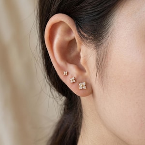May include: Three gold stud earrings arranged in a row along the earlobe. Each earring features a four-petaled flower design composed of small, clear, round-cut stones set in a gold metal base, with each petal being a single stone.