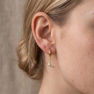 May include: A gold hoop earring featuring a crescent moon charm. The charm is embellished with a small, iridescent round opal stone at the centre and tiny clear pavé crystals along the curved edge of the moon, suspended from a thin gold hoop.