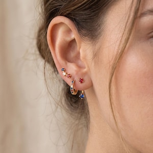 May include: A set of gold earrings featuring a small hoop with alternating blue and red stones, a three-stone ear climber with blue, white, and red gems, and a single red stud earring, plus a dangling blue stone earring attached to the hoop.