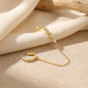 May include: A gold-tone ear cuff connected by a delicate chain to a stud earring. The ear cuff features a row of small clear stones, while the stud component consists of four clear stones arranged in a line, all crafted in a polished metal finish.
