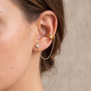 Op de afbeelding: Een goudkleurige ear cuff met drie horizontale ribbels, verbonden door een fijn gouden kettinkje aan een enkele ronde oorsteker met een heldere steen. Het sieraad heeft een gepolijste metalen afwerking en een schitterende zetting.