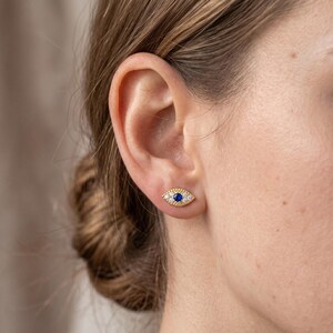 May include: A gold stud earring in the shape of an eye, featuring a central round blue stone surrounded by small clear stones set in a textured gold frame. The earring is positioned on the earlobe, showcasing a detailed, symmetrical design.
