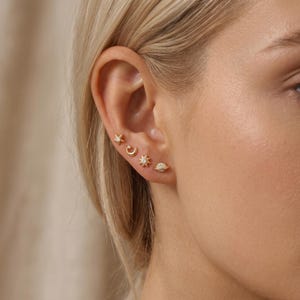 May include: A set of four small gold stud earrings arranged in a row along the earlobe. The designs include a star, a crescent moon, a sunburst, and a planet, each featuring tiny clear stones that catch the light for a subtle, shimmering effect.