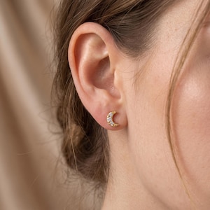 May include: A gold-toned stud earring shaped like a crescent moon. The earring features a textured, beaded edge and is set with three small, clear, round-cut stones along the curve of the moon, secured in the earlobe with a post backing.