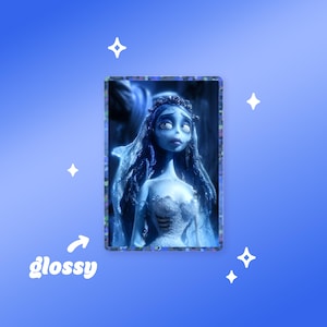 May include: A rectangular sticker featuring a blue-toned illustration of a character with long dark hair, a veil, and a white dress. The sticker has a dark, speckled border and a glossy finish. The word glossy is printed in white with an arrow pointing to it.