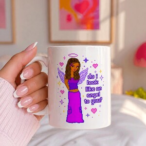 May include: White ceramic mug featuring a pixelated illustration of an angel with brown hair, purple outfit, and text that reads "do i look like an angel to you?". The design includes hearts and stars.