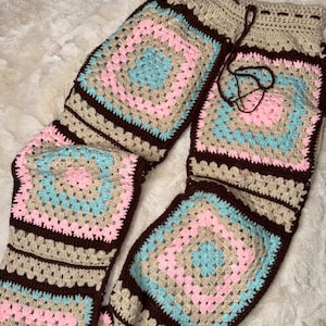 Pode incluir: Calças de crochê feitas à mão com um padrão quadrado em cores pastel. As calças têm cintura com cordão e são feitas com fio marrom, bege, rosa e azul. O design de crochê é visível em toda a calça.